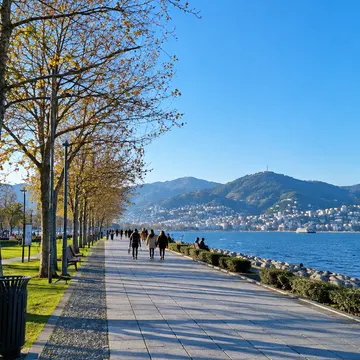People walking on a paved waterfront path under autumn trees with a blue sea and mountain city in the distance