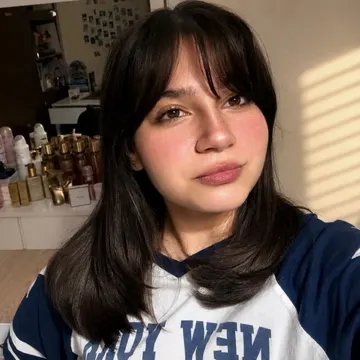 Young woman with dark hair wearing a blue and white shirt in a sunlit room with perfume bottles on a shelf behind her