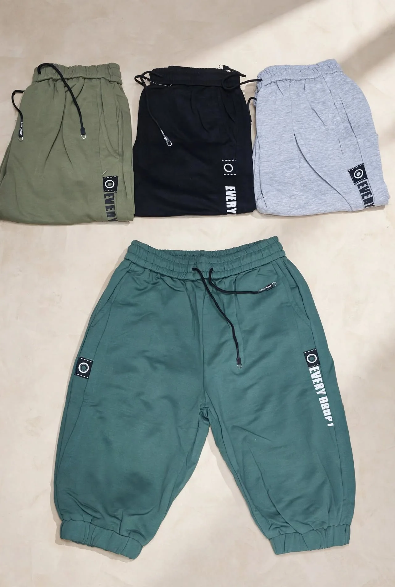 Four pairs of drawstring sweatpants in olive black gray and teal laid flat on a light beige surface