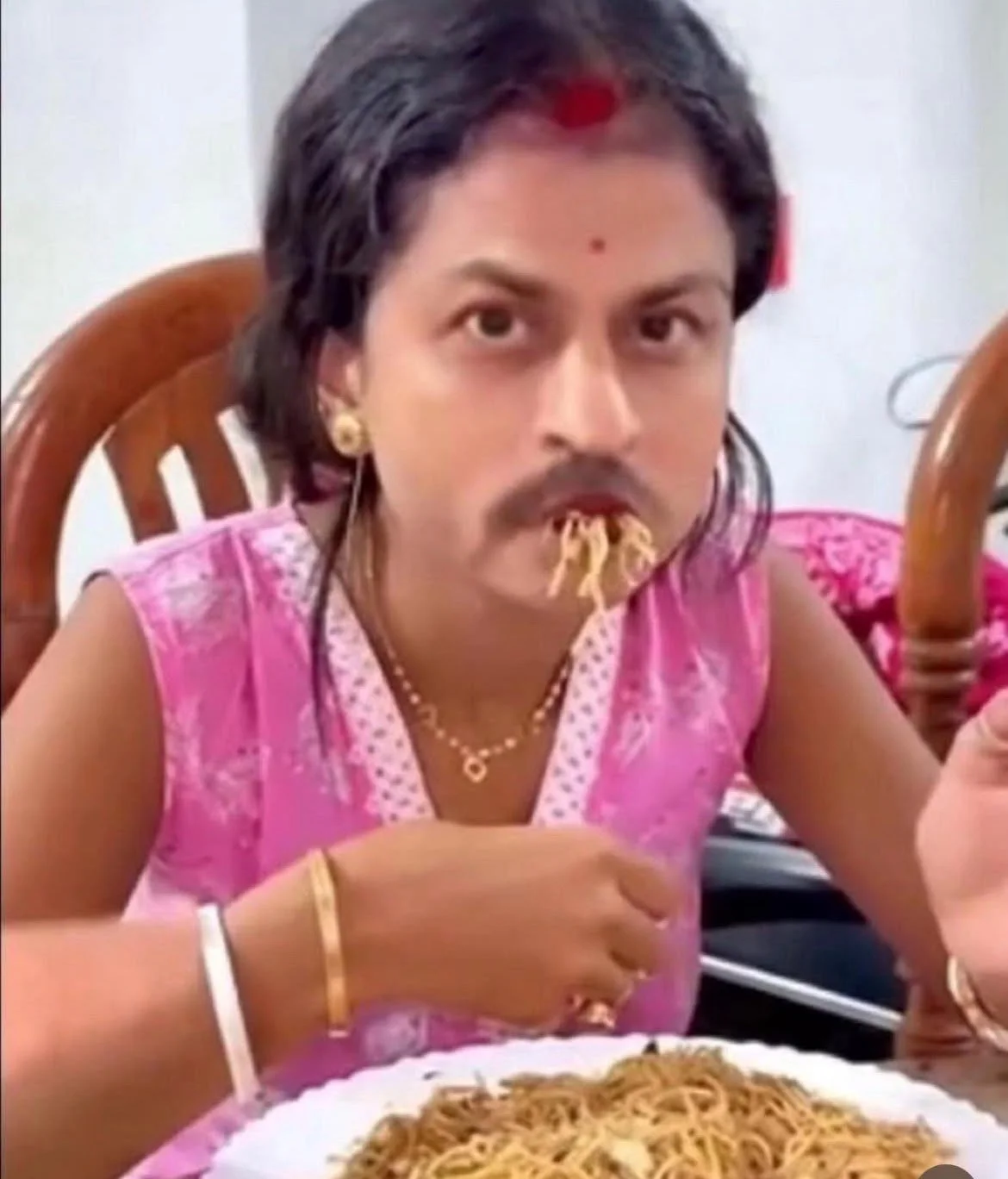 Person wearing pink top and gold jewelry eating noodles with noodles in mouth