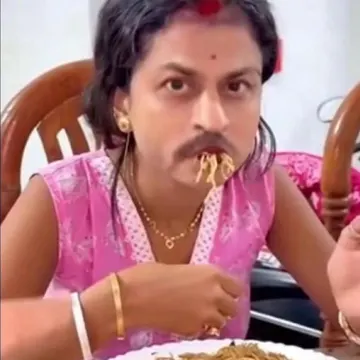 Person wearing pink top and gold jewelry eating noodles with noodles in mouth