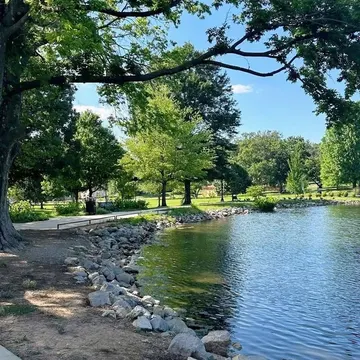 A paved path winds along a rocky lakeshore shaded by leafy trees under a bright blue sky