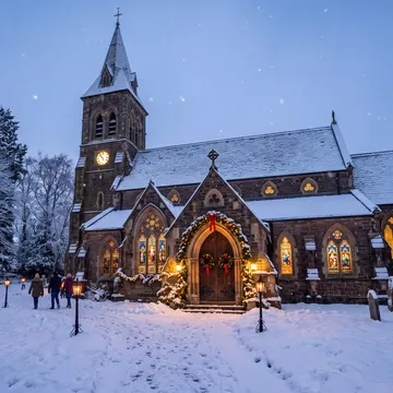 A snow-covered stone church with a steeple and illuminated windows, surrounded by snow-laden trees and lampposts, with a few people walking in the for