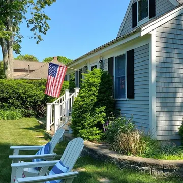 Two white adirondack chairs with blue cushions on a grassy lawn near a house with an american flag