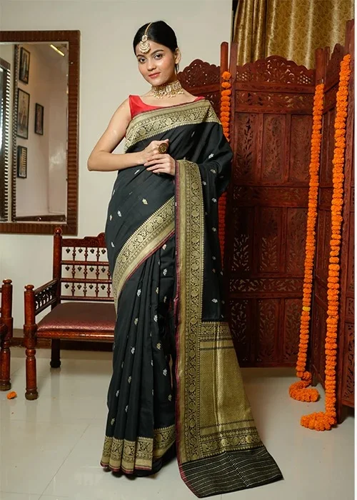 Woman wearing black and gold embroidered saree with red blouse standing near wooden screen with marigold garlands