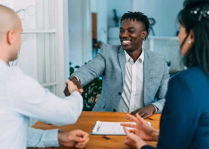 Three people in business attire shaking hands over a wooden table with documents