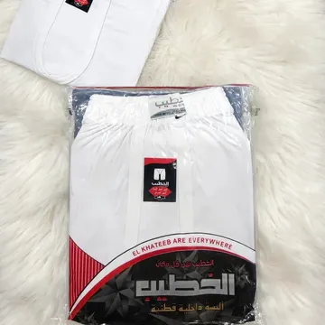 Two white El Khateeb shorts one folded one packaged on fluffy white rug with TIRAZ plaque