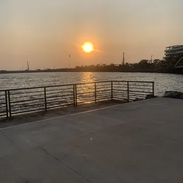 A sunset over a body of water viewed from a concrete pier with a metal railing, with buildings and industrial structures visible in the distance