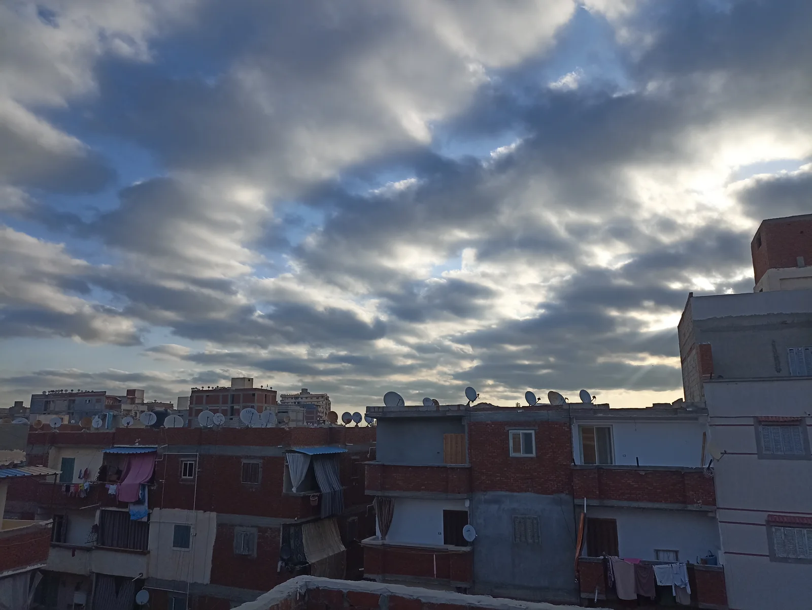 Urban rooftops with satellite dishes and laundry under a dramatic cloudy sky at dusk