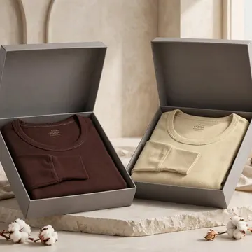 Two folded ribbed shirts in brown and beige in gray boxes with cotton bolls and dried grasses on stone