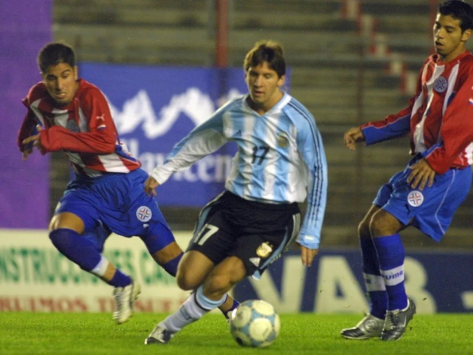 Soccer player in light blue and white jersey dribbling ball with two opponents in red and white jerseys chasing
