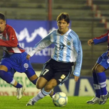 Soccer player in light blue and white jersey dribbling ball with two opponents in red and white jerseys chasing