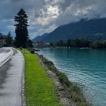 A paved path runs beside a grassy shore along a turquoise lake with mountains and clouds in the background