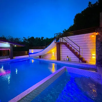 A brightly lit pool with a slide and stairs at night surrounded by trees and stone walls
