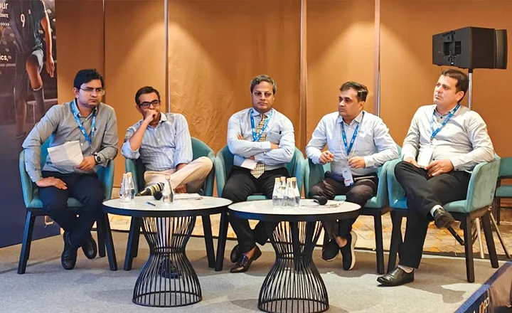 Five men in business attire seated at a conference panel with microphones and water bottles