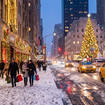 A snowy city street at dusk with pedestrians walking past decorated storefronts and yellow taxis driving on a wet road, featuring a large illuminated