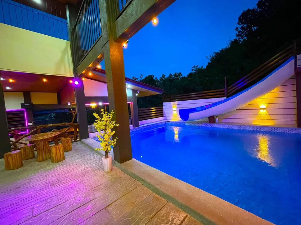A lit pool area at dusk with a slide and wooden seating under string lights