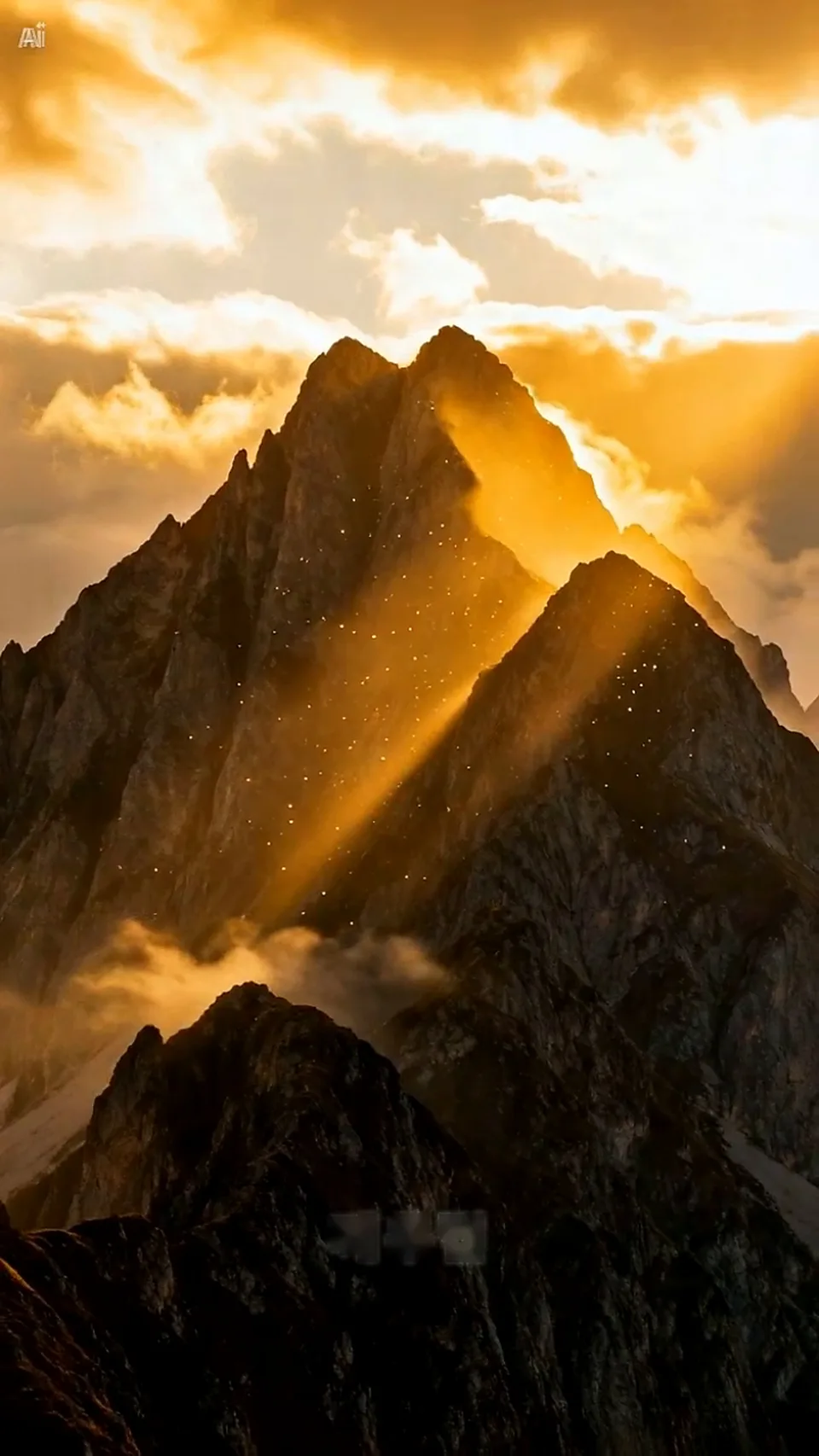 A mountain peak is illuminated by golden sunlight breaking through clouds, with a faint watermark visible in the lower center