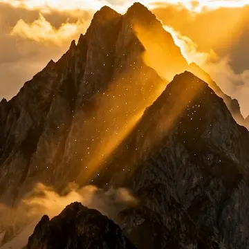 A mountain peak is illuminated by golden sunlight breaking through clouds, with a faint watermark visible in the lower center